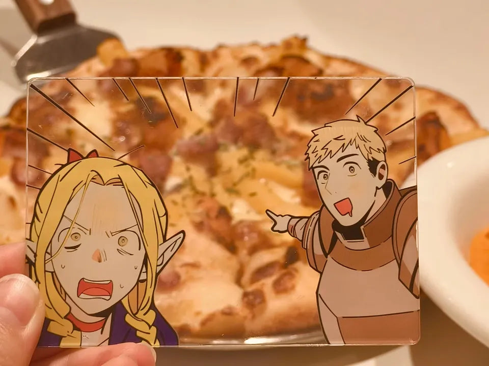 An acrylic transparent card featuring Delicious in Dungeon (Dungeon Meshi) characters Marcille and Laios in a playful food-themed scene. Marcille is wide-eyed with shock, while Laios grins excitedly, pointing to the center of the image with drool dripping from his mouth—as if spotting an irresistible meal. The card is propped against a backdrop of delicious real-world dish (pizza), blending the anime’s fantasy hunger with mouthwatering reality. The translucent acrylic gives 